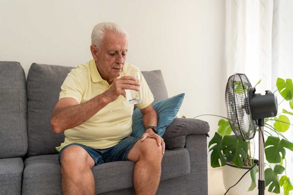senior,man,drinking,water,on,sofa,with,electric,fan,cooling