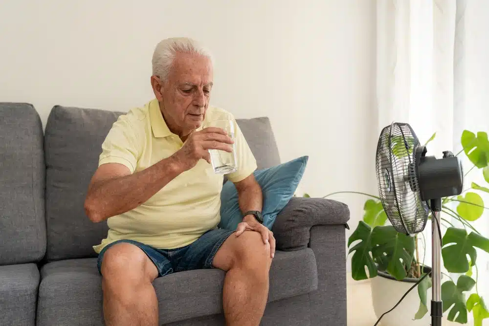 senior,man,drinking,water,on,sofa,with,electric,fan,cooling