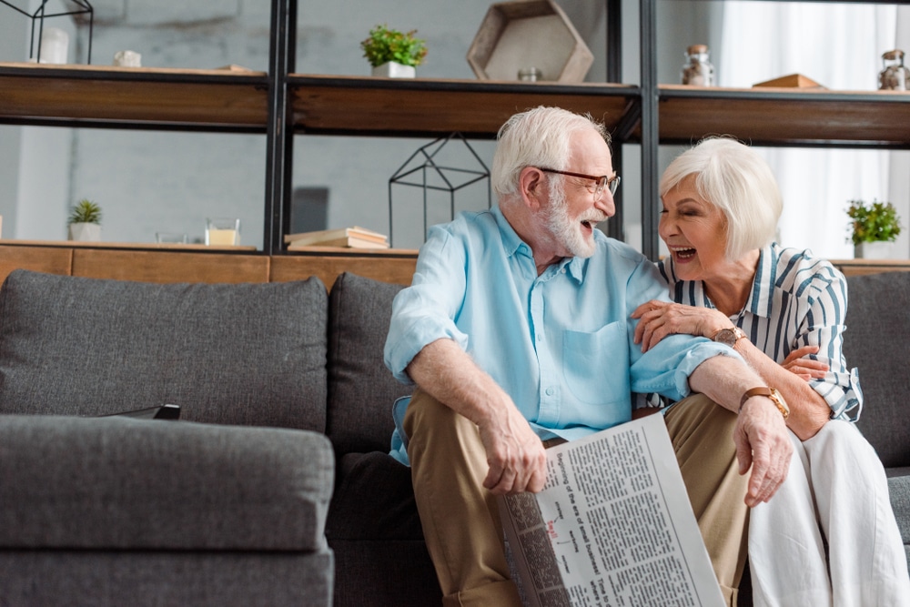 senior,couple,with,newspaper,laughing,while,sitting,on,couch,on