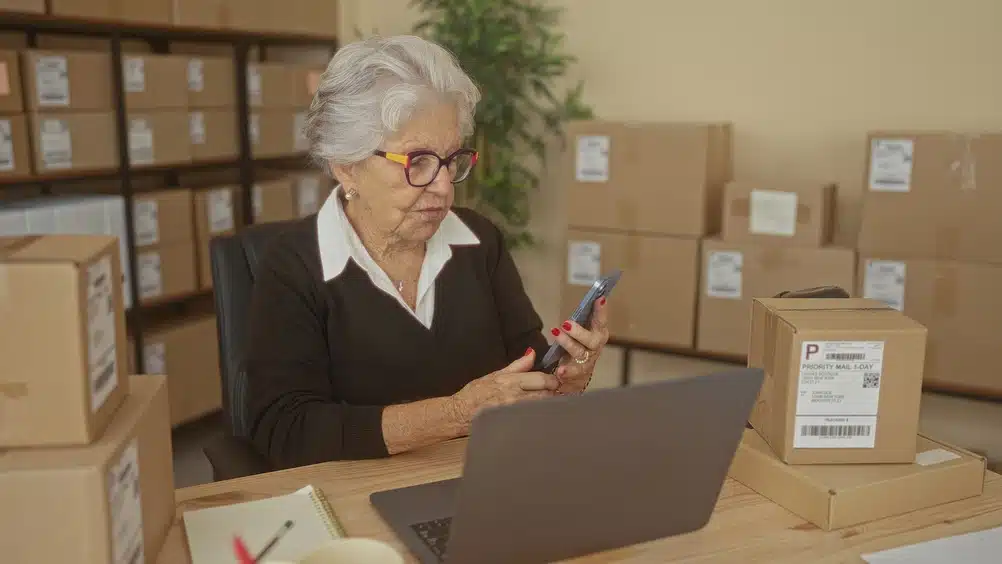 elderly,woman,typing,on,laptop,and,tapping,smartphone,among,stacked